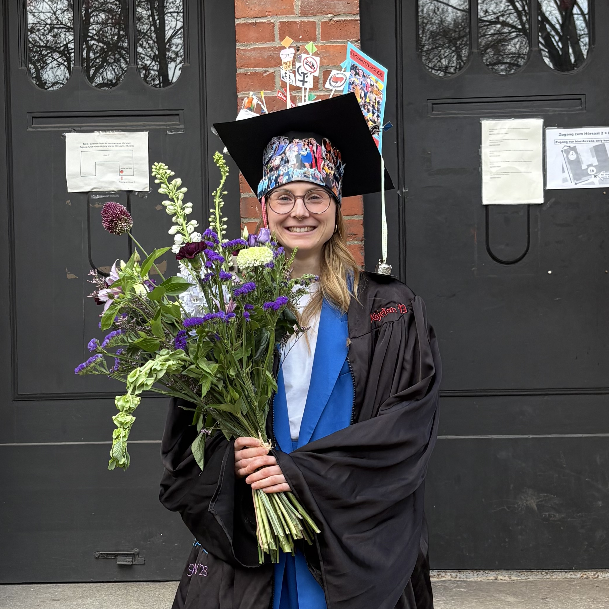 Viola Hollek standing infront of a red brick building, wearing doctoral cap, robe, and a colorful flower bouquet